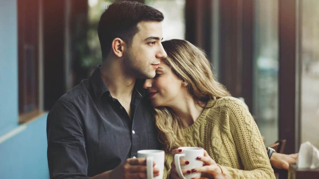 A man and woman sit close together, each holding a white mug, in a cafe.