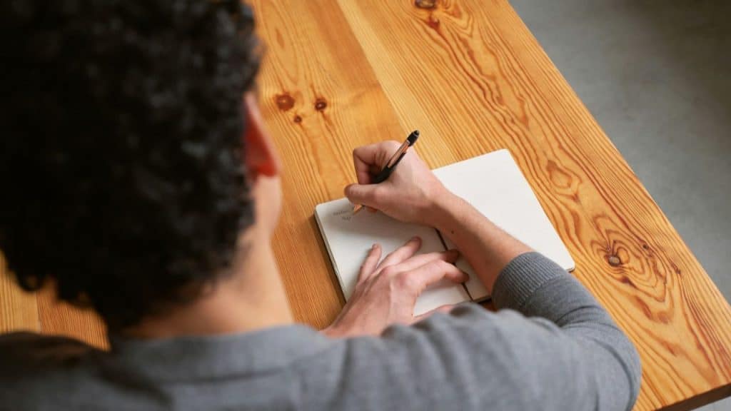 A person with curly hair is seen from behind, writing in an open notebook on a wooden table.