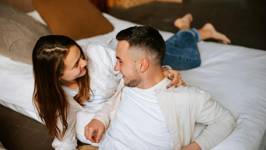 A smiling couple looks at each other on a bed, with the woman's arm around the man.