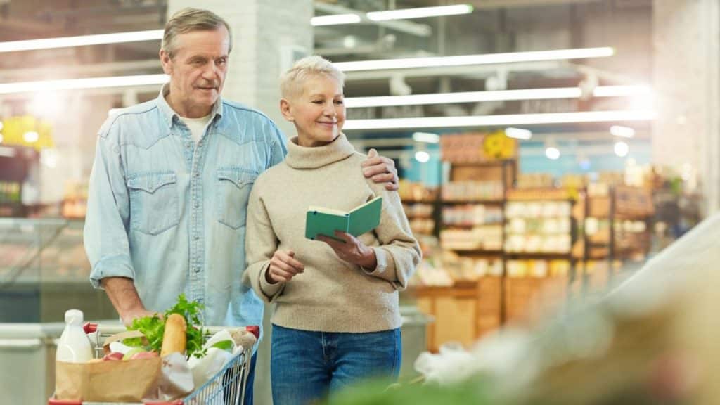 An elderly couple is grocery shopping, with the man pushing a cart and the woman holding a small book.