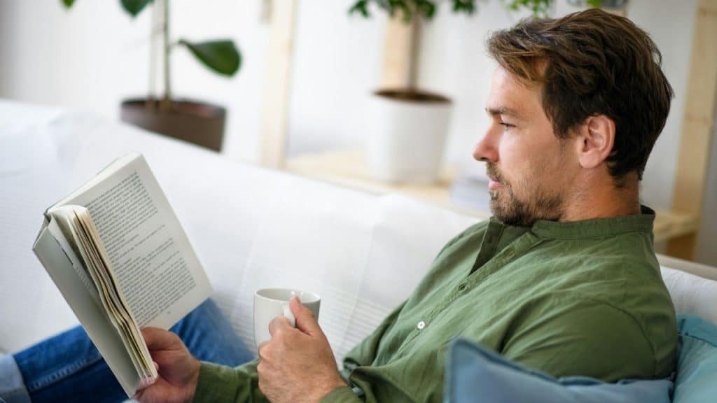 A man with a beard holds a book and a mug, sitting on a couch.