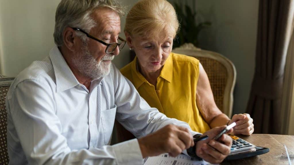 An elderly couple looks intently at a mobile phone, a calculator nearby.