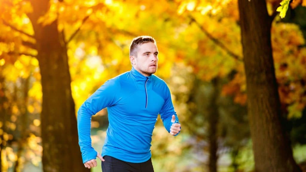 A man in a blue long-sleeved shirt is jogging outdoors in a park with autumn leaves.