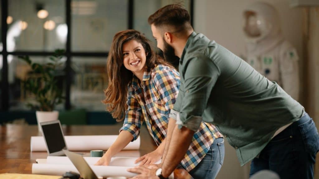 A smiling woman in a plaid shirt looks at a man who is leaning over a table with rolled papers.