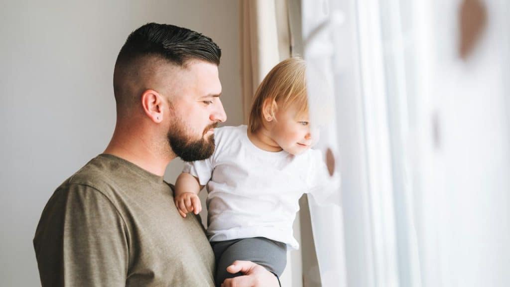 A bearded father holding his toddler as they look out a window.