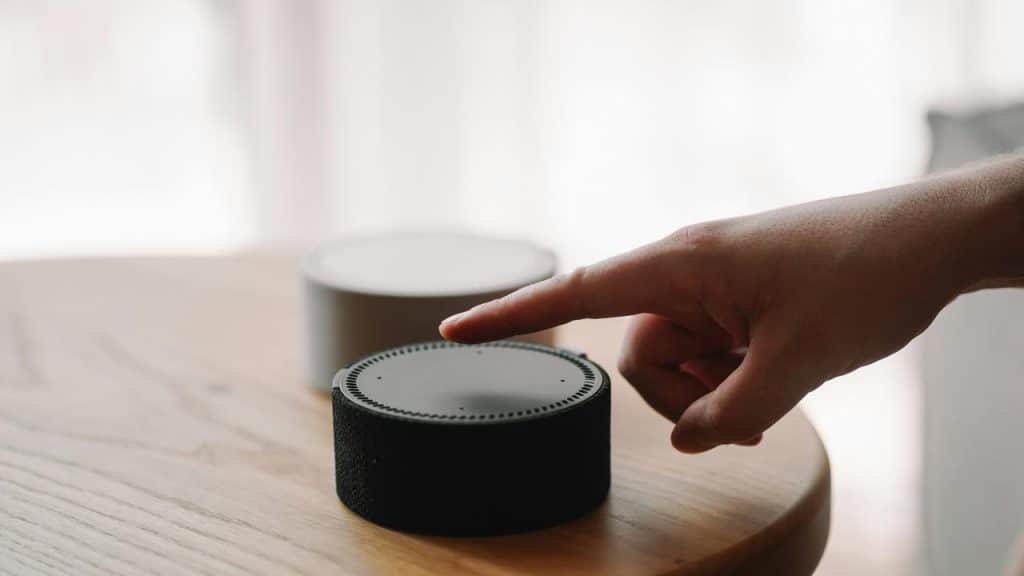 A hand reaching to tap a smart speaker device on a wooden table, with another speaker in the background.