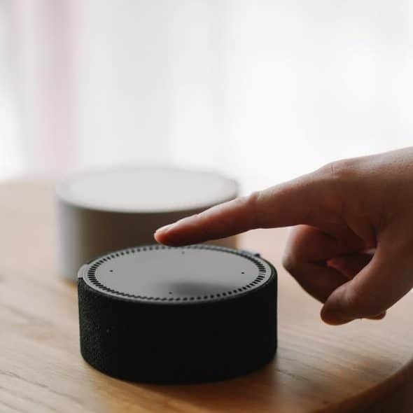 A hand reaching to tap a smart speaker device on a wooden table, with another speaker in the background.