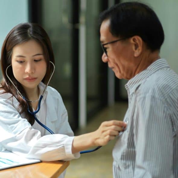 A female doctor using a stethoscope to examine an older male patient in a clinical setting.