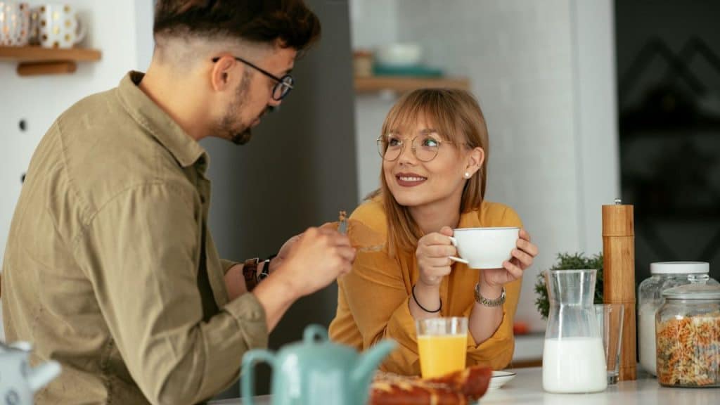 A smiling woman with glasses holds a cup and looks at a man across the table.