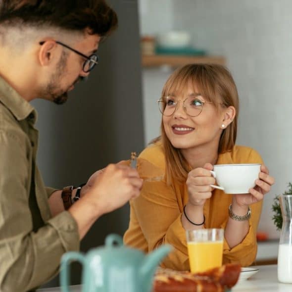 A smiling woman with glasses holds a cup and looks at a man across the table.