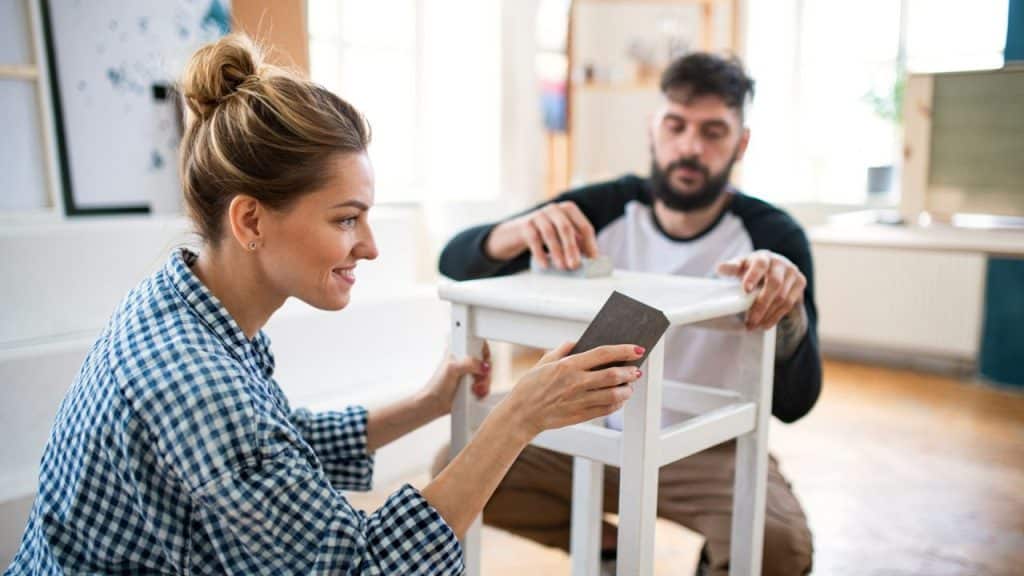 A smiling woman sands a white stool while a man works on another in the background.