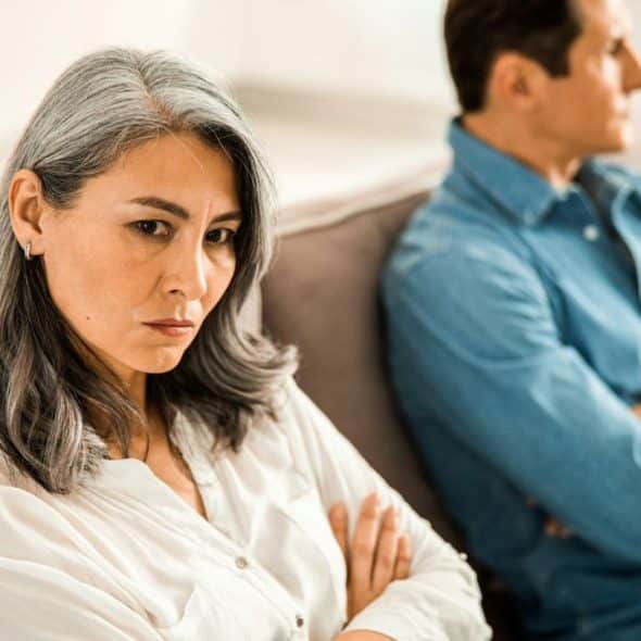 A woman with crossed arms looks annoyed, while a man faces away on a sofa.