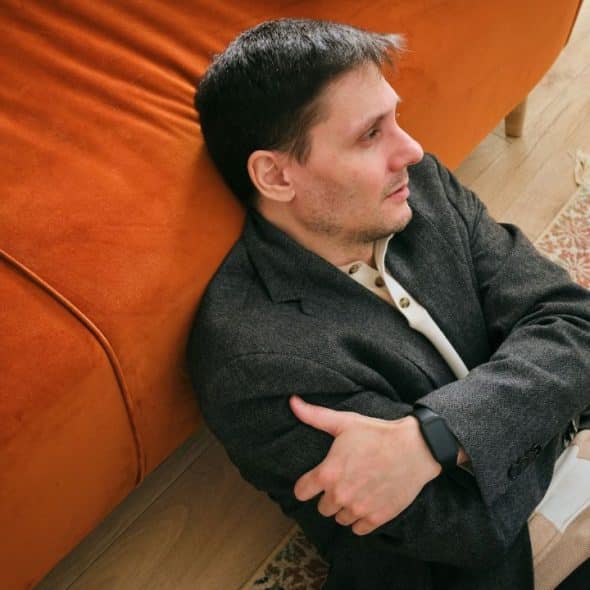 A man in a jacket sits on the floor with his arms crossed, leaning against a couch.