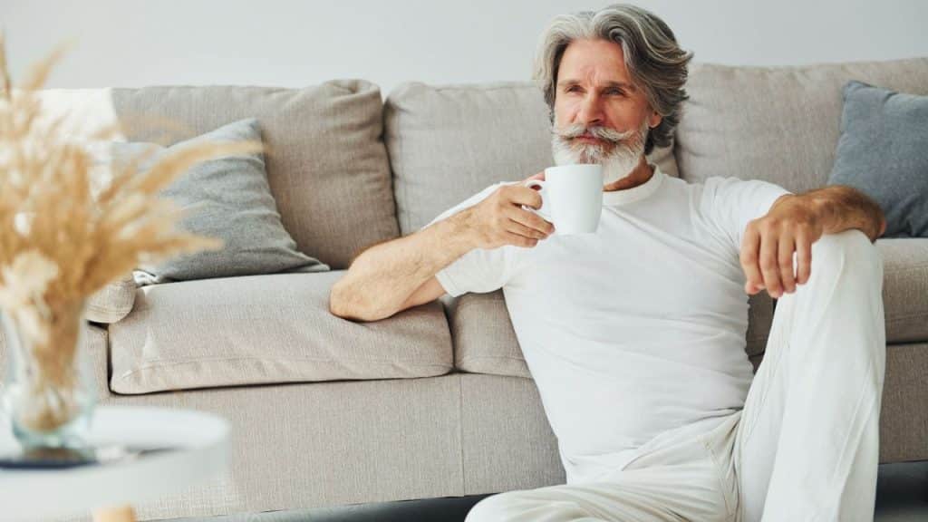 A distinguished man with a white beard and hair sits by a couch, holding a mug.