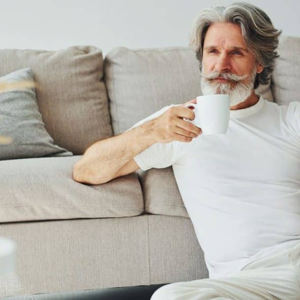 A distinguished man with a white beard and hair sits by a couch, holding a mug.