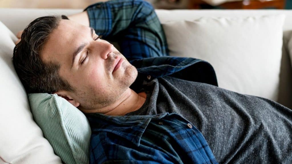 A man in a plaid shirt and gray tee naps peacefully on a white couch.