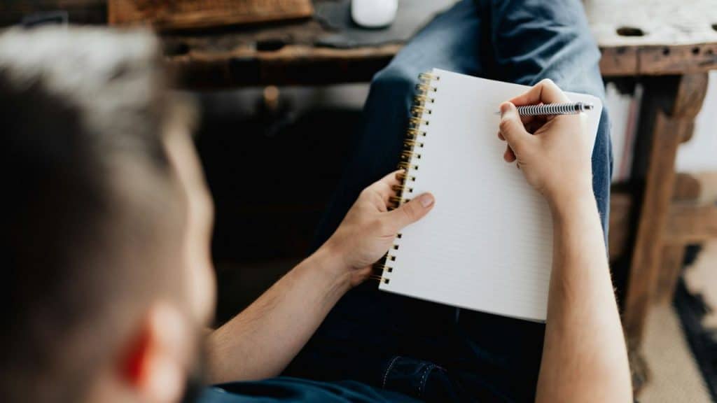 A person, viewed from behind, writes in a spiral-bound notebook on their lap.