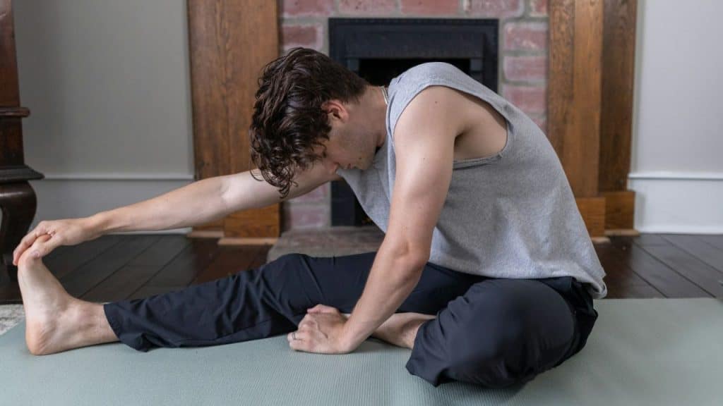 A man in a grey tank top and dark pants stretches on a mat, reaching for his foot.
