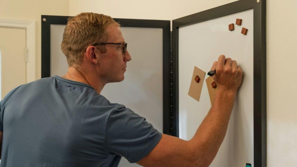 A man in glasses writes on a whiteboard with attached brown paper and wooden magnets.