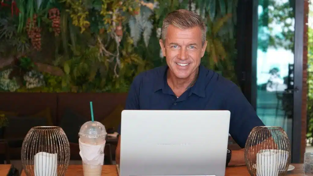 A smiling man with graying hair sits at a table with a laptop and a drink.