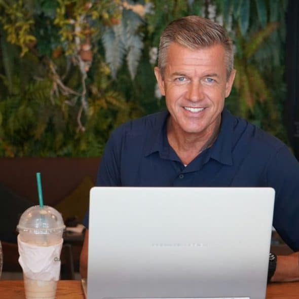 A smiling man with graying hair sits at a table with a laptop and a drink.