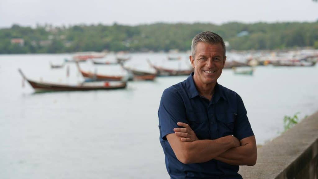 A smiling man with crossed arms stands by a body of water with boats in the background.
