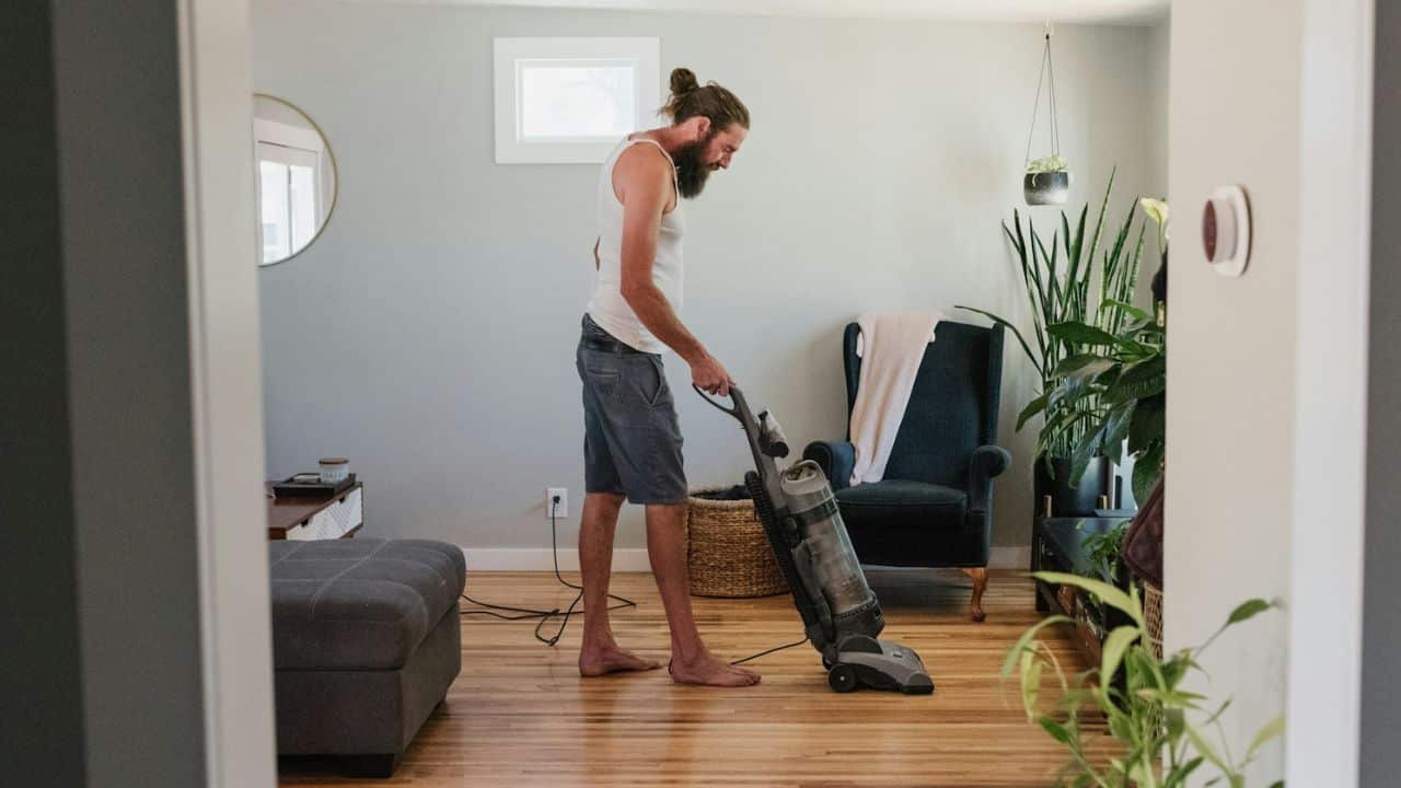 A bearded man with hair in a bun vacuums a wooden floor in a living room.