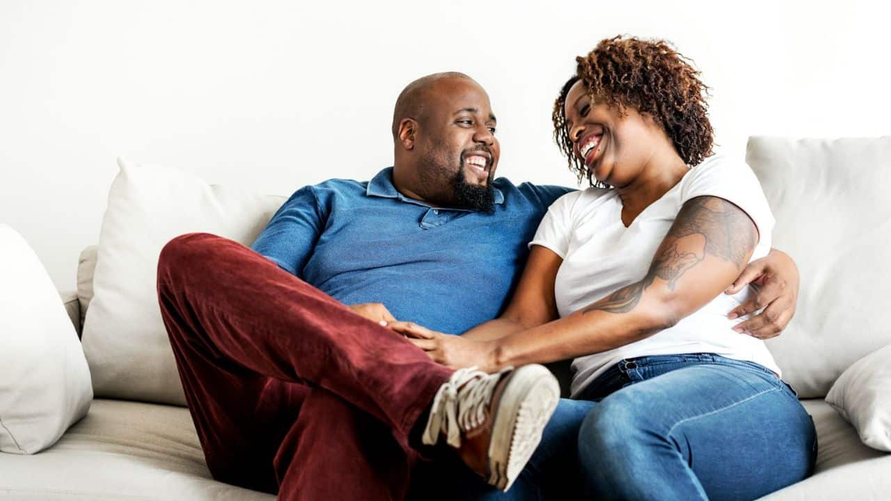 A couple sits on a white couch, smiling and looking at each other.