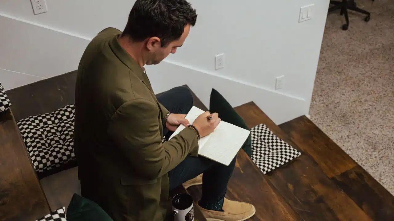 A man in a blazer sits on wooden stairs, writing in a notebook.