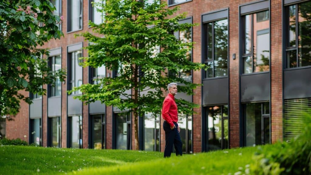 A man in a red shirt walks on a grassy area in front of a brick building.