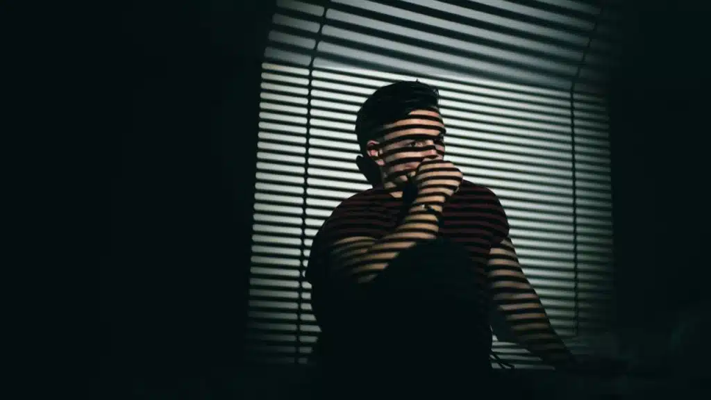 A man with short hair sits in a dark room with light stripes from blinds on him.