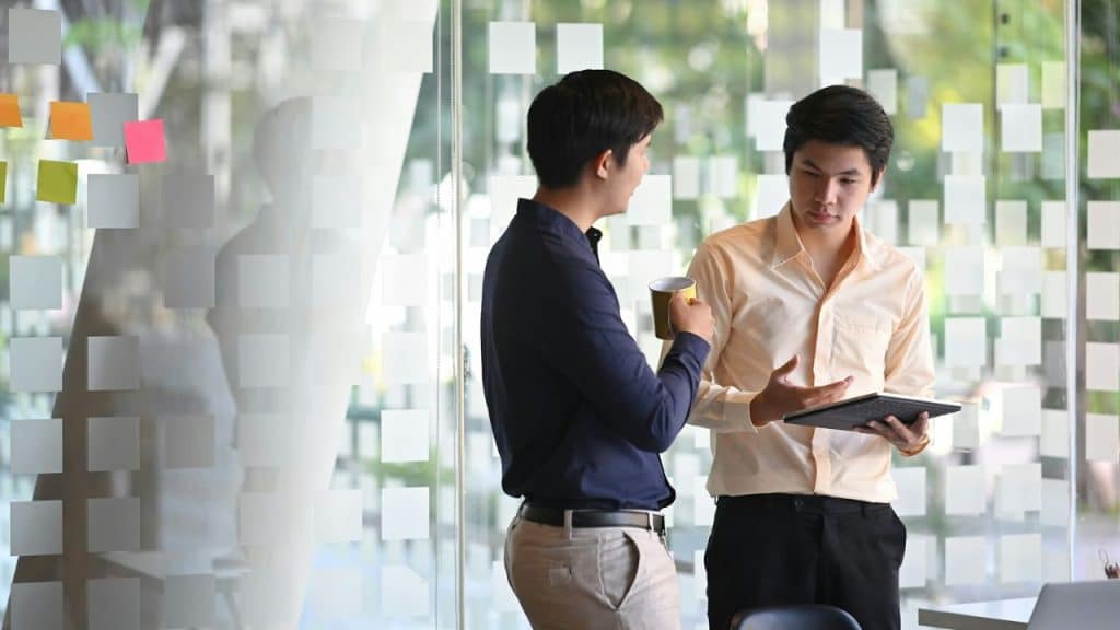 Two men are talking; one holds a yellow mug, the other a tablet, in front of a glass wall with sticky notes.