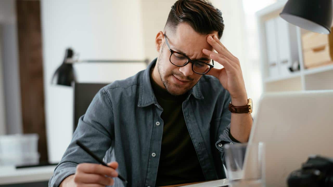A frustrated man holding his head while working on a laptop.