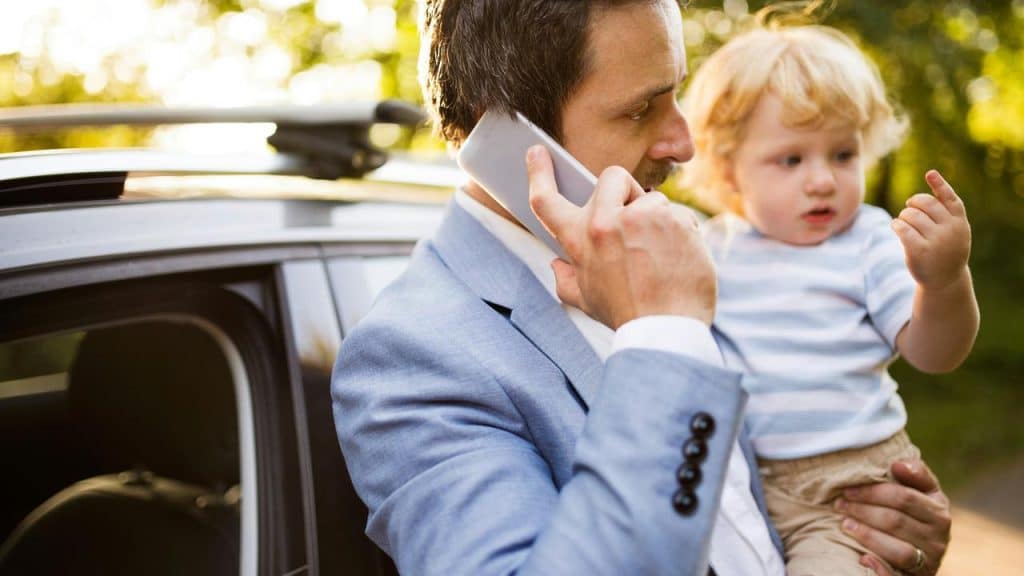 A man holding a toddler while talking on his phone beside a car.
