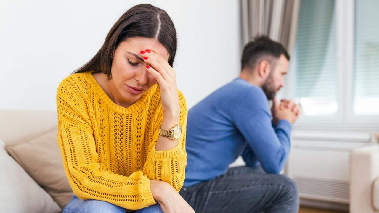 A distressed woman sits in the foreground with a man looking away in the background.