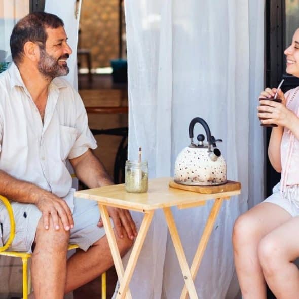 A man and a woman sit at a small table outdoors, conversing and smiling.