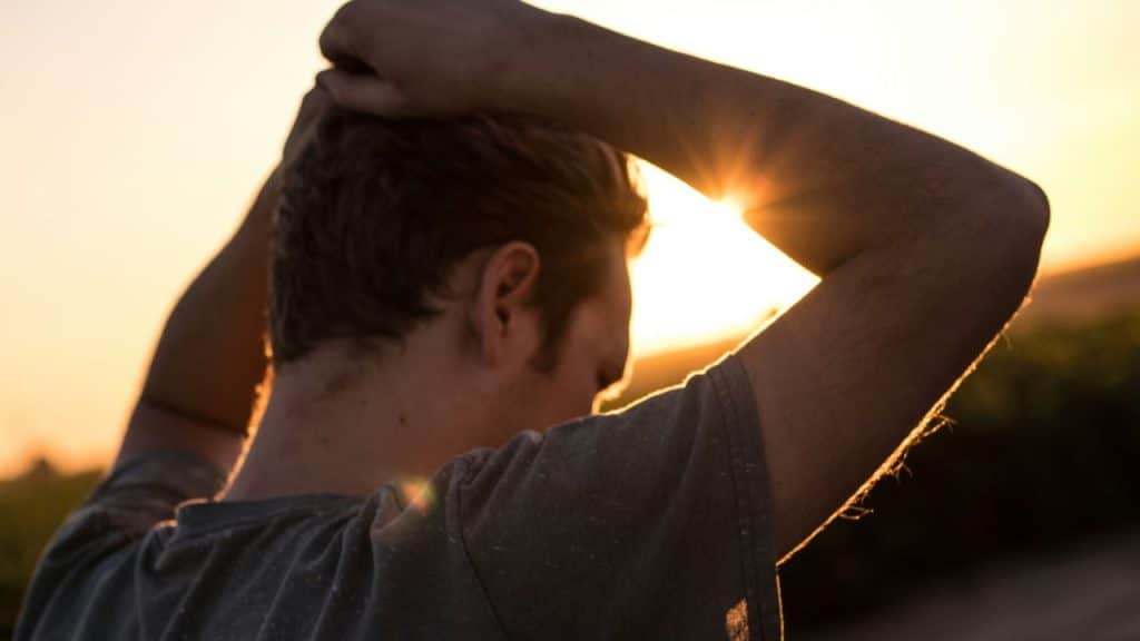 A person from behind, with hands on their head, facing a bright, setting sun.