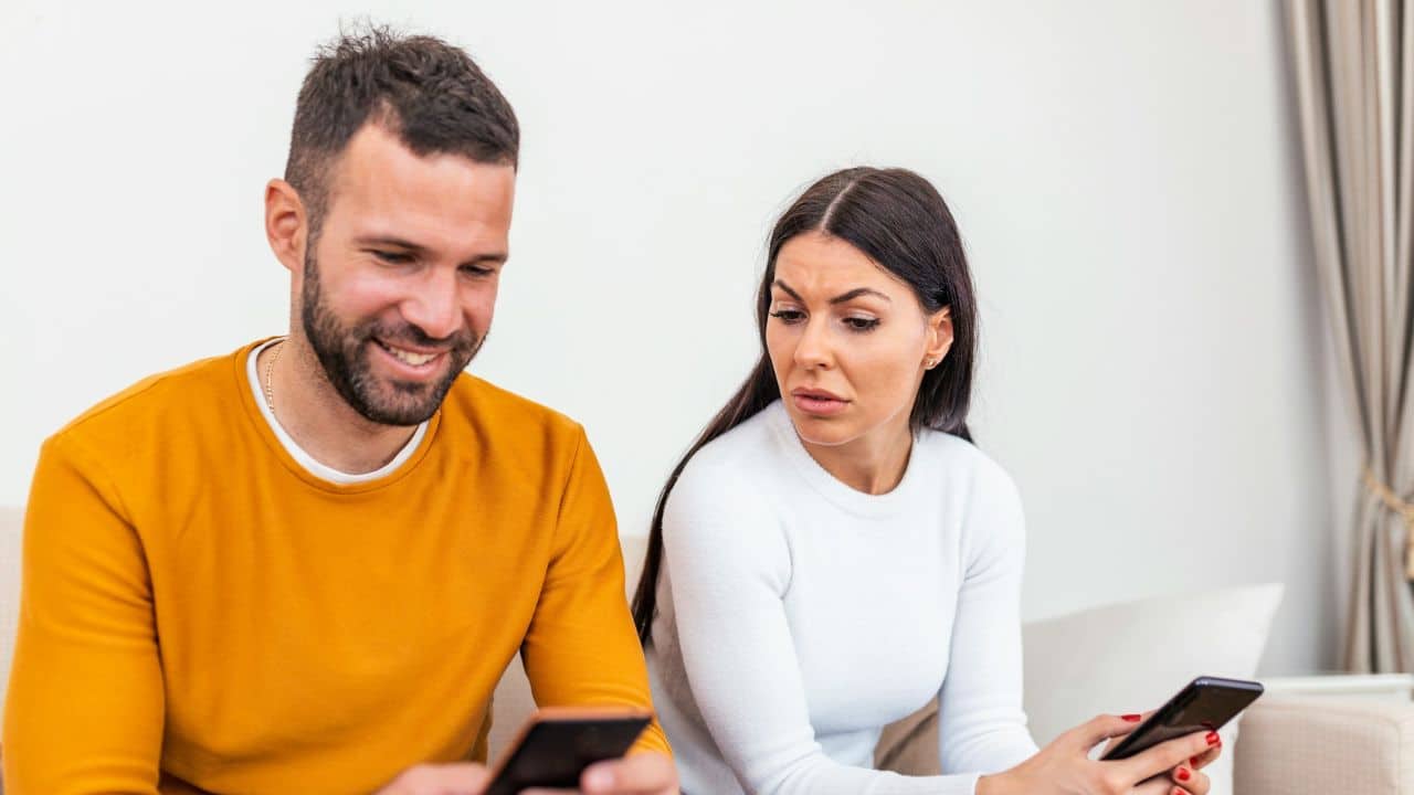 A man smiles while looking at his phone, as a woman beside him looks unhappily at hers.