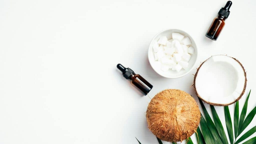 A flat lay of whole and halved coconuts, a bowl of coconut chunks, and amber dropper bottles.