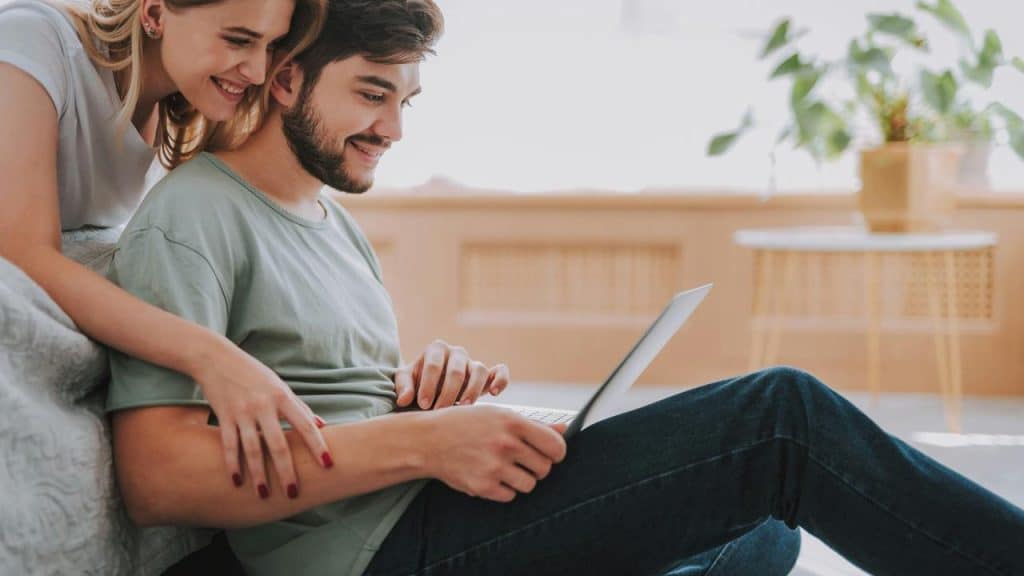 A smiling couple leaning on a sofa while the man works on a laptop.