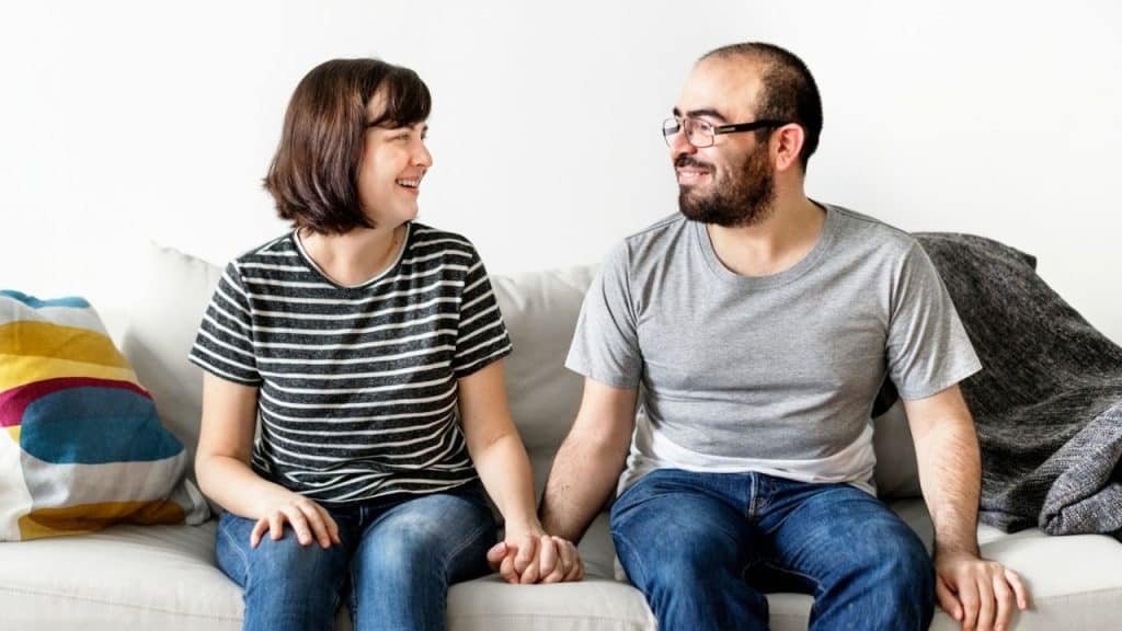 A smiling couple sits on a couch, holding hands and looking at each other.