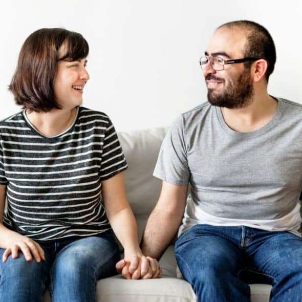 A smiling couple sits on a couch, holding hands and looking at each other.