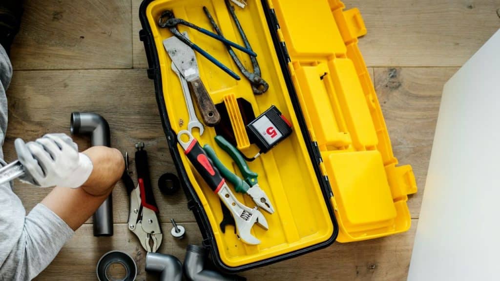 A top-down view of a yellow open toolbox filled with various tools, next to pipe fittings.