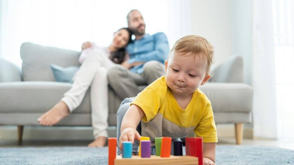 A baby in a yellow shirt plays with a wooden toy on the floor, while adults sit on a couch in the background.