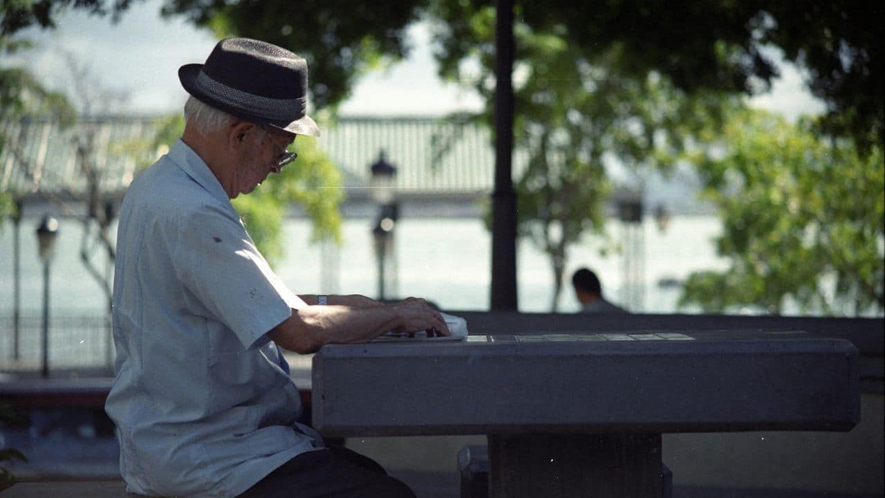 A man in a fedora sits at an outdoor table, looking down at something..