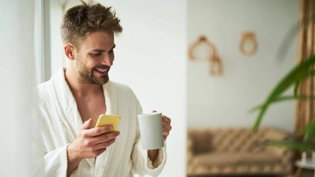 A smiling man in a white robe holds a phone and a mug.