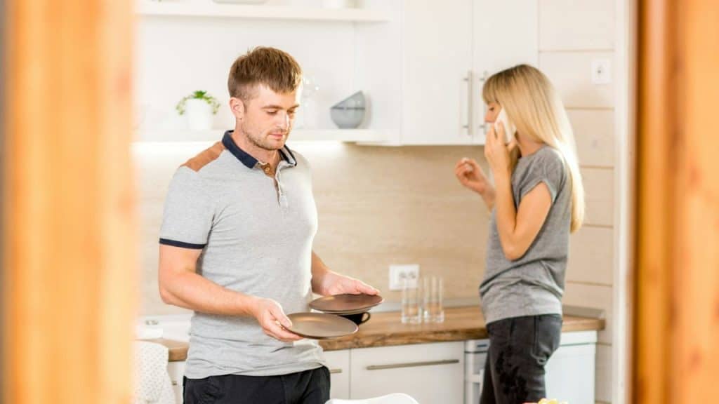 A man holds two plates while a woman talks on the phone in a modern kitchen.