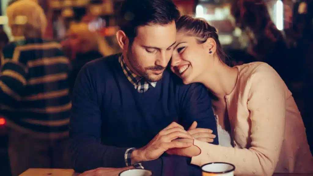 A woman leans into a man at a bar, both smiling and holding hands.