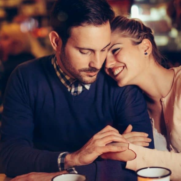 A woman leans into a man at a bar, both smiling and holding hands.