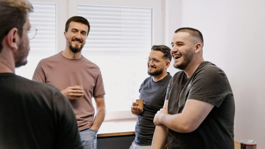 Four men are gathered together, smiling and laughing, with two holding cups.